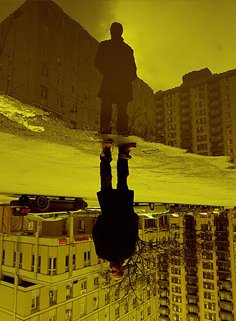 Split screen, full length portrait of Danilo Deluxo standing in front of an apartment block. In the top portion of the frame he is silhouetted; in bottom half of the frame he is upside down like a reflection.